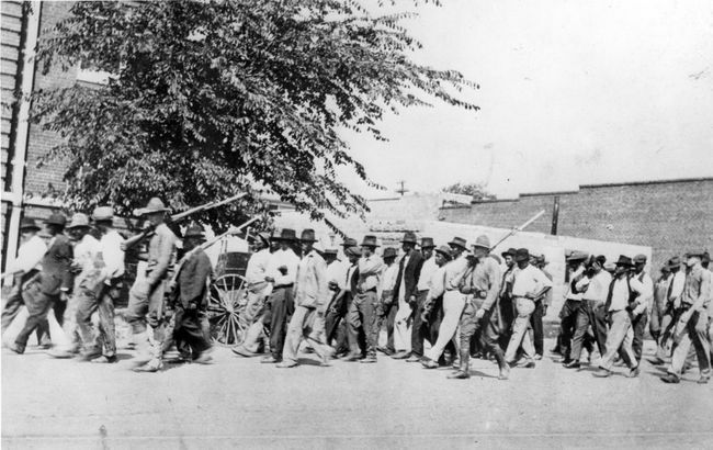 קבוצה של חיילים של המשמר הלאומי, נושאת רובים עם כידונים מחוברים, מלוות גברים שחורים לא חמושים למרכז מעצר לאחר הטבח במירוץ טולסה, טולסה, אוקלהומה, יוני 1921.
