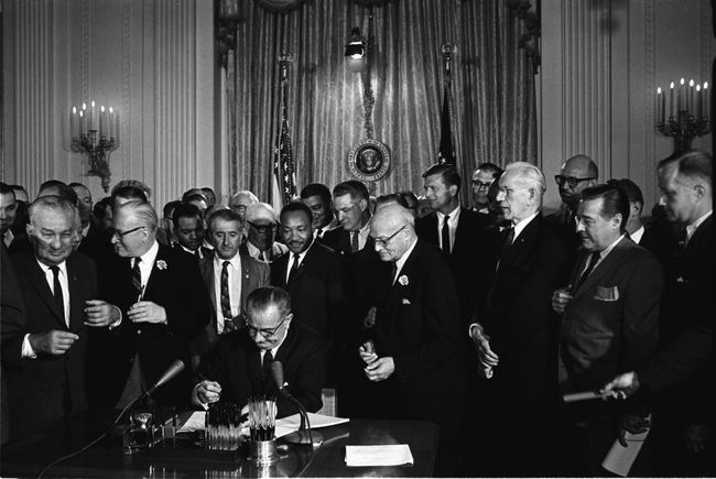 הנשיא לינדון ב. ג'ונסון חותם על חוק זכויות האזרח משנת 1964 כשמרטין לותר קינג, ג'וניור ואחרים, מסתכלים.