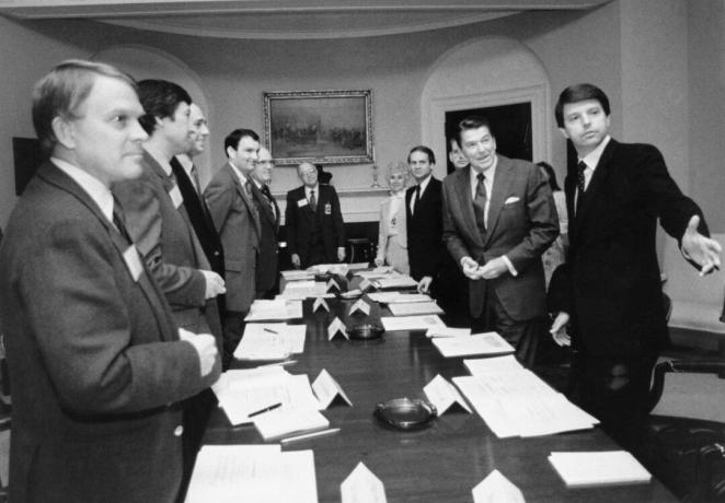 תמונה בשחור לבן של רונלד רייגן ועוד כמה גברים בחליפות סביב שולחן ישיבות ארוך